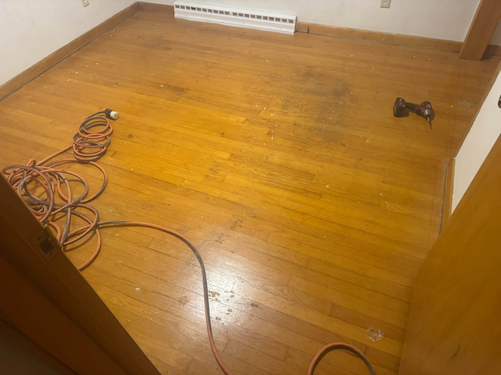 Worn hardwood floor before refinishing, with a cord and a drill on the boards