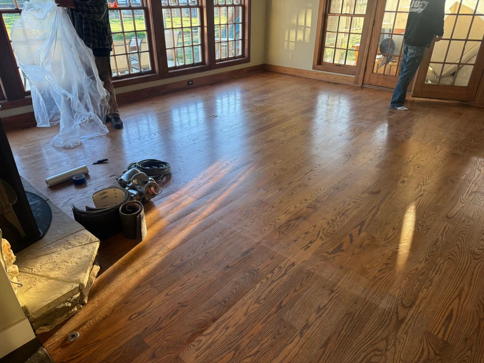 Lighter hardwood floor in a room with wood windows and French doors, with a sander, sanding belts, and plastic sheeting still in the room